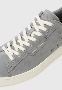 Szürke velúr sneakers fehér cipőfűzőkkel, finom varrással, megerősített orral és dombornyomott logóval az oldalon. Texturált gumitalp.