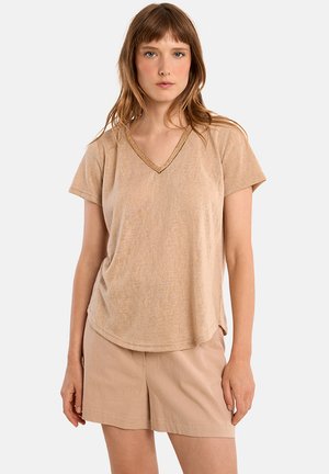 Donna con capelli castano chiaro che indossa una maglietta beige a maniche corte con scollo a V e pantaloncini beige, in piedi davanti a uno sfondo chiaro uniforme.