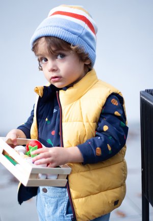 Enfant portant un gilet rembourré jaune par-dessus une chemise marine avec des imprimés de légumes, tenant une caisse en bois de fruits et légumes en jouet. Bonnet rayé bleu et rouge.