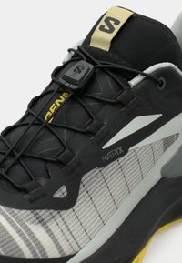 Μαύρο και γκρι αθλητικό παπούτσι με επάνω μέρος από δίχτυ, καουτσούκ σόλα και αντανακλαστικές λεπτομέρειες. Διαθέτει στρογγυλεμένη μύτη, σύστημα κορδονιών και μπρανταρισμένες πινελιές.