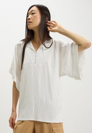 Blusa bianca ampia con scollatura a V, maniche corte a campana, tessuto testurizzato e due bottoni frontali, abbinata a pantaloni corti beige.