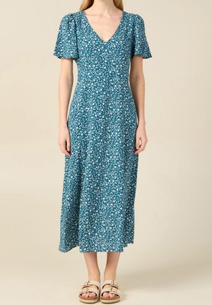 Nainen pukeutuneena tealin väriseen midi-mittaiseen mekkoon, jossa valkoinen kukkakuosi, lyhyet hihat ja V-aukko, yhdistettynä beigen värisiin sandaaleihin neutraalia taustaa vasten.