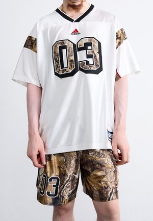 Person, die ein weißes Adidas-Trikot mit Camouflage-"03" und Ärmeln sowie passende Camouflage-Shorts mit "03" am linken Bein trägt, steht still.