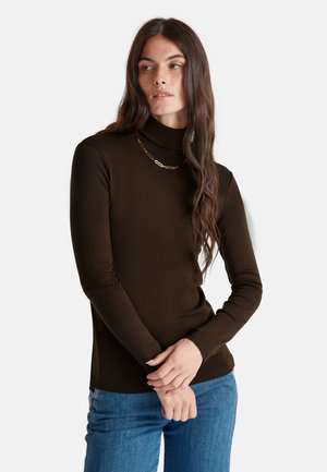 Vrouw met lang golvend bruin haar, gekleed in een donkerbruine coltrui en een gouden ketting, poseert met haar handen gevouwen voor zich.