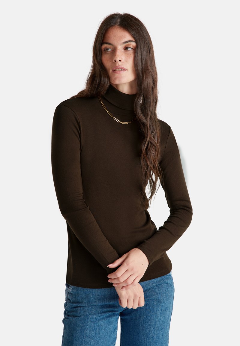 Frau mit langen, welligen braunen Haaren, die einen dunkelbraunen Rollkragenpullover und eine goldene Halskette trägt, posiert mit gefalteten Händen vor der Brust.