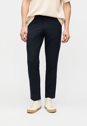 Mann, der schwarze, schmal geschnittene Hosen, ein beiges T-Shirt und cremeweiße Sneaker mit Gummisohlen trägt, steht vor einem schlichten Hintergrund.