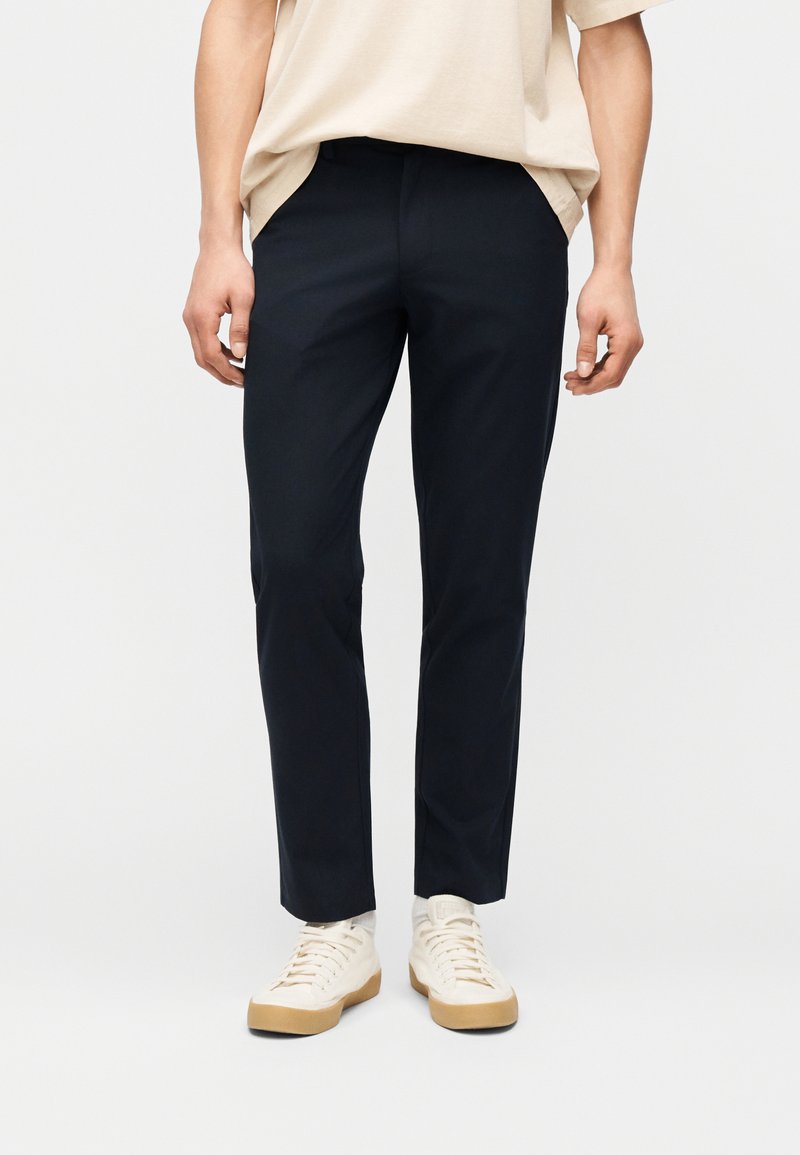 Άνδρας που φοράει μαύρο παντελόνι στενής εφαρμογής, μπεζ μπλουζάκι και εκρού αθλητικά παπούτσια με καουτσούκ σόλες, στέκεται μπροστά σε ένα απλό φόντο.