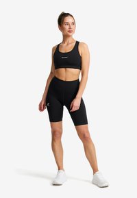 Kvinna stående i svart sportbh och cykelbyxor med vita sneakers, ser åt sidan mot en vit bakgrund.