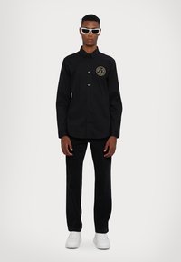 Camicetta nera con bottoni e patch logo, abbinata a pantaloni neri e sneakers bianche. Il modello indossa occhiali da sole bianchi, in piedi davanti a uno sfondo neutro.