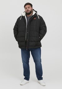 Sort dunjakke med en hvid fleeceforet hætte, featuring en frontlynlås, knapper og et brunt logo patch, parret med blå jeans og hvide sneakers.