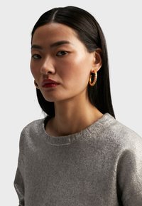 Silver, texturerad tröja med rund halsringning. Modellen bär stora guldörhängen. Håret är rakt och långt, och ramar in ansiktet.