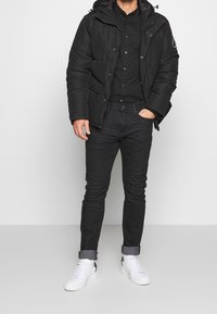 Czarna kurtka puchowa z kapturem, zapinana na guziki, z kieszenią na piersi, połączona z ciemnymi dżinsami i bia�łymi trampkami. Gładka tekstura, nowoczesny design.