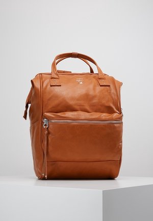 Zaino in pelle marrone con tasca frontale con zip, doppie maniglie superiori e branding minimalista, posizionato su una superficie bianca contro uno sfondo semplice.