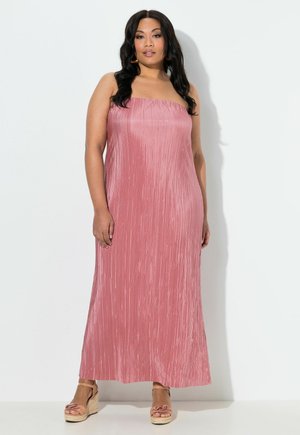 Vrouw met lang donker haar, gekleed in een strapless, geplooide roze maxi-jurk en beige sleehakken, staand tegen een witte achtergrond.