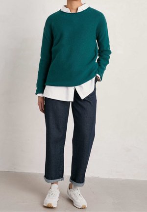 Personne portant un pull sarcelle par-dessus une chemise blanche, un jean foncé retroussé et des baskets blanches, debout contre un mur et un sol clairs et unis.
