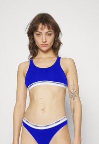 Blå sports-bh og matchende truse, laget av teksturert stoff. Begge har en kontrastfarge hvit midje med "TOMMY JEANS"-logo.