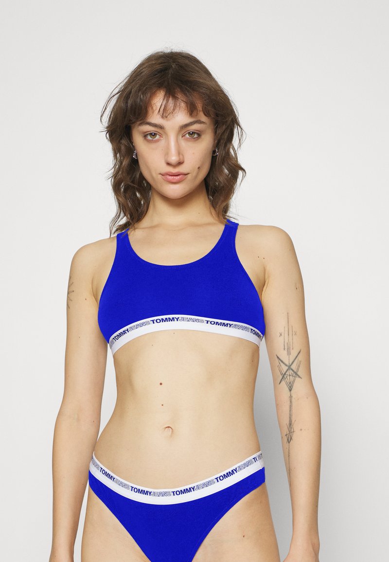 Blå sports-bh og matchende truse, laget av teksturert stoff. Begge har en kontrastfarge hvit midje med "TOMMY JEANS"-logo.