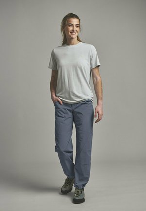 Lächelnde Frau mit langen Haaren, die ein hellgraues T-Shirt, blaue Hosen und Wanderschuhe trägt, steht mit einer Hand in der Tasche.