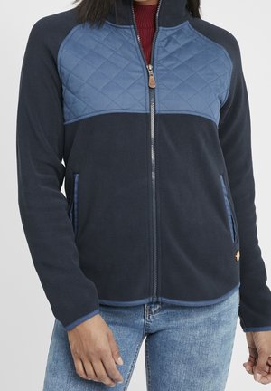 Kvinde iført en marineblå fleecejakke med lynlås og quiltet øvre brystpanel samt lyseblå jeans.