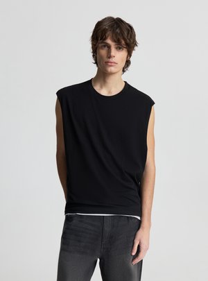 Giovane con capelli castani di media lunghezza, indossa una canottiera nera e jeans scuri, in piedi davanti a uno sfondo chiaro e uniforme.