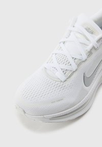 Gros plan d'une chaussure de running Nike blanche avec tige en mesh, lacets blancs et logo Swoosh argenté sur fond clair.