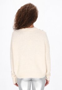 Crème-kleurige, losgebreide trui met een zachte textuur, geribbelde manchetten en decoratieve knopen op de schouders. Draagt met glanzende zilveren broek.