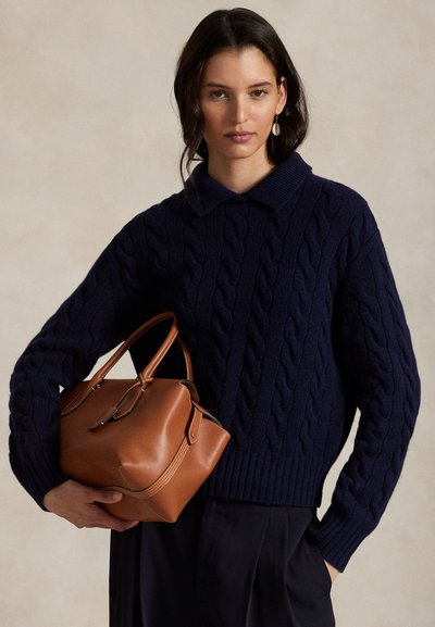 Frau trägt einen marineblauen Zopfpullover und hält eine mittelbraune Ledertasche vor einem schlichten beigen Hintergrund.