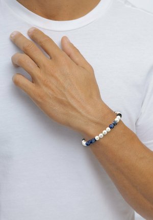 Armband av marinblå och vita pärlor med silverdetaljer, med ett pärlat mönster och slitstark snodd, bärs på en solbränd hand.