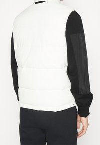 Weißer Steppwesten mit einem gesteppten Design und schwarzen Strickärmeln. Verfügt über einen hohen Kragen und eine glatte, matte Oberfläche. Keine sichtbaren Elemente.