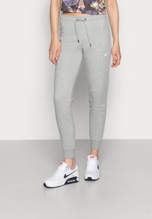 Kvinde iført lysegrå Nike joggingbukser med snøre og hvide Nike Air Max-sneakers, stående på en glat grå gulv.