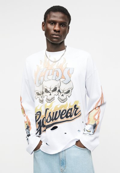 Άνδρας με λευκό πουκάμισο μακρυμάνικο με σχέδια κρανίων, φλόγες και γάτα, φορεμένο με ανοιχτόχρωμο μπλε τζιν, στέκεται μπροστά σε απλό φόντο.