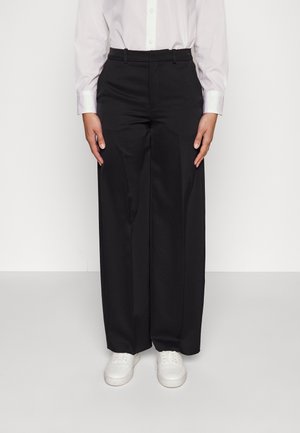 Person iført sorte brede bukser, hvid skjorte med knapper og hvide sneakers, der står mod en ensfarvet baggrund.