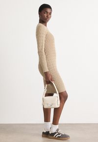 Stickad beige klänning med flätmönster, långa ärmar och ribbad fåll. Matchad med grå sneakers och en liten vit handväska.
