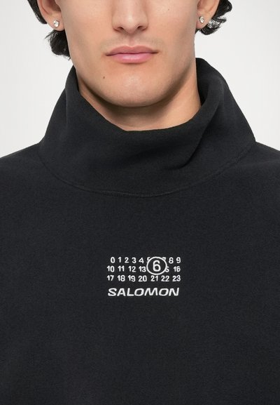 Fekete gyapjú garbó fehér grafikai nyomattal. Kerek szám design és "SALOMON" logó található a mellkason. Puha, texturált anyag.