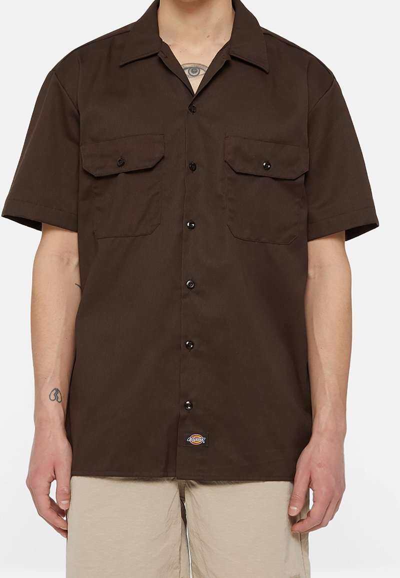 Muž v tmavohnědém košilovém triku s krátkým rukávem, které má dvě náprsní kapsy, béžových šortkách a s viditelnými tetováními na krku a předloktí.