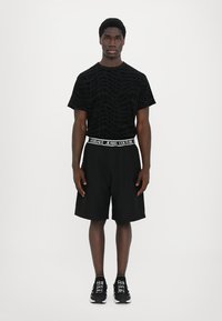 Juodas trumpas rankovėmis marškinėlis su tekstūruotu logotipo raštu, derinamas su juodomis šortais, turinčiais Versace Jeans Couture prekės ženklo juosmenį. Juodi sportbačiai.