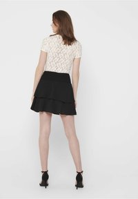 Spitzenoberteil mit kurzen Ärmeln in Creme mit transparentem Rücken, kombiniert mit einem schwarzen, mehrlagigen Rüschenshorts und schwarzen High-Heel-Sandalen.