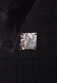 Zwarte gebreide stof met een zilveren vierkante hardwaredetail met een gegraveerd gezichtsontwerp, aangevuld met een donker fluwelen accent.