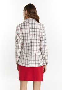 Blazer à carreaux blancs avec des motifs rouges et navy, en tissu texturé, présente des manches longues et des accents de boutons dorés. Porté sur une jupe rouge.