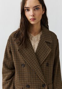 Braun karierter Doppelreiher-Mantel mit großen Revers, schwarzen Knöpfen und lockerer Passform, über einem beigen Pullover.