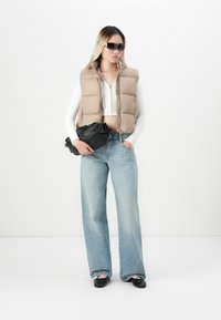 Kvinne iført beige puffervest, hvit kort topp, vide jeans, svarte leiligheter og solbriller, holder en svart håndveske mot en hvit bakgrunn.