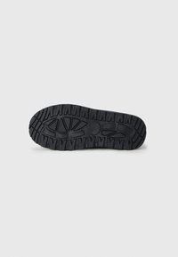 Semelle de chaussure en caoutchouc noir avec un motif de semelle robuste, présentant de profondes rainures et un accent circulaire au centre.