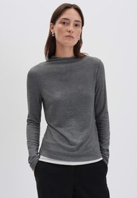 Frau in einem enganliegenden, grauen Langarmoberteil, die mit beiden Händen in den Taschen einer schwarzen Hose steht, vor einem schlichten, hellen Hintergrund.