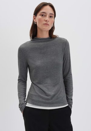 Donna che indossa un top grigio a maniche lunghe aderente con le mani nelle tasche di pantaloni neri, in piedi contro uno sfondo chiaro uniforme.