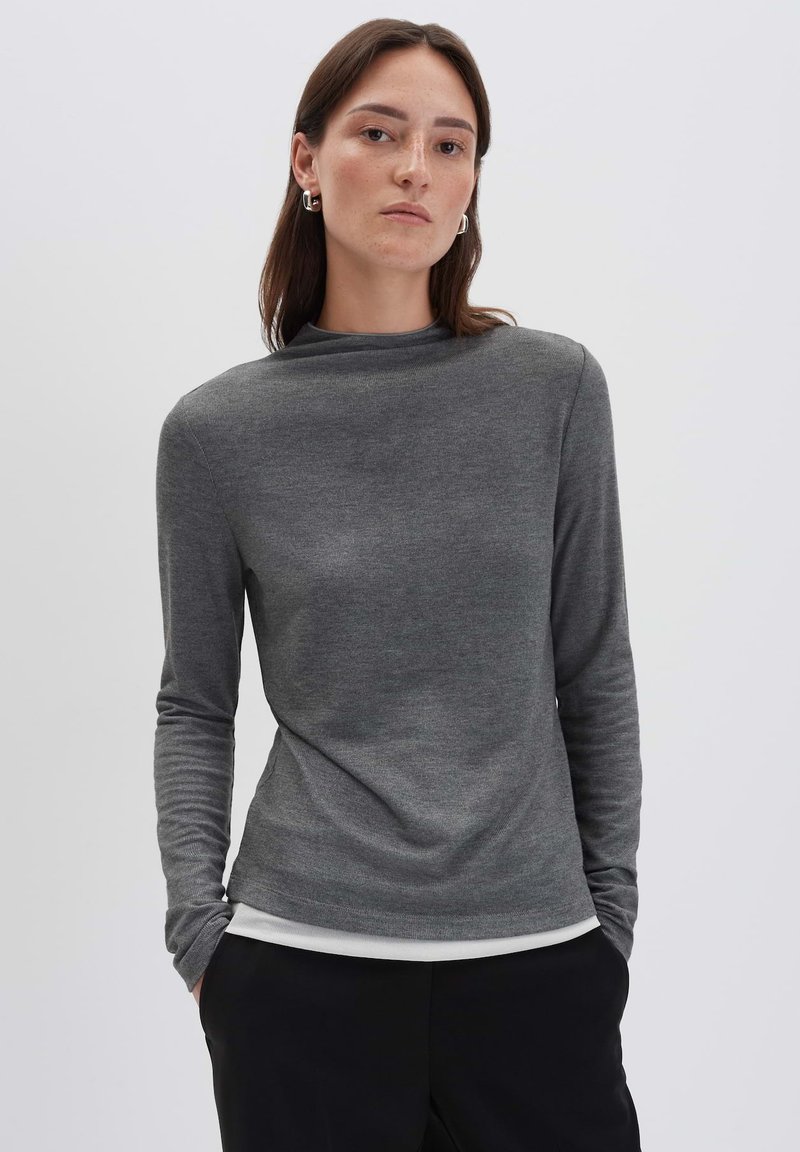 Frau in einem enganliegenden, grauen Langarmoberteil, die mit beiden Händen in den Taschen einer schwarzen Hose steht, vor einem schlichten, hellen Hintergrund.