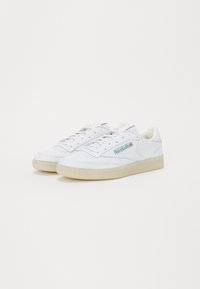 Λευκά αθλητικά παπούτσια με λείο επάνω μέρος από δέρμα, διάτρητο μέρος στα δάχτυλα και λαστιχένια σόλα. Διαθέτουν πράσινο λογότυπο στο πλάι και κορδόνια.