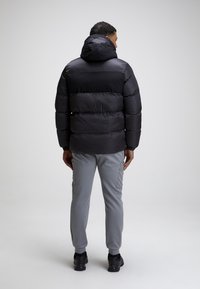 Giacca a puffo nera con cappuccio, caratterizzata da sezioni a cuciture orizzontali. Indossata con pantaloni grigi a gamba attillata e scarpe nere, vista da dietro.