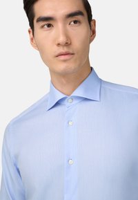 Svetlo modra srajca z gumbi, izdelana iz teksturiranega materiala, s klasičnim ovratnikom in preprostimi belimi gumbi, ki ima prilegajoč se kroj in ozke rokave.