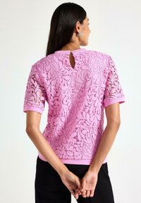 Nainen, jolla on pitkät tummat hiukset, pukeutuneena vaaleanpunaiseen pitsitoppiin, jossa on kukkakuviota, seisoo kädet selän takana.