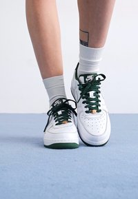 Beli usnjeni športni copati z zelenimi poudarki in vezalkami, ki imajo perforiran prednji del in zlate detajle. Noseni so s svetlo sivo nogavico.